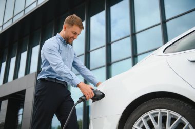 Gayri resmi giyinmiş genç bir adamın portresi, şarj istasyonunda duruyor. Eko elektrikli araba konsepti
