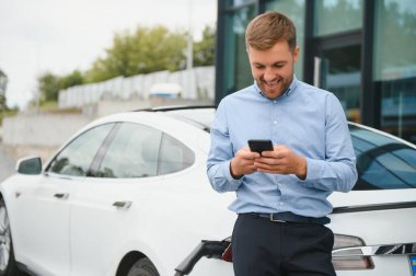 Handsome man in business suit surfing internet on modern smartphone while waiting electric car to charge