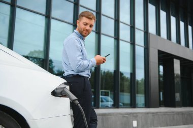 Man charging his electric car at charge station and using smartphone.