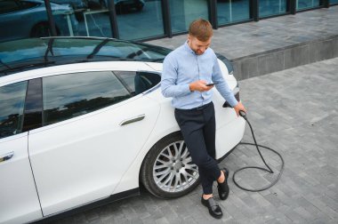 portrait of young handsome bearded man in casual wear, standing at the charging station and holding a plug of the charger for an electric car. Eco electric car concept