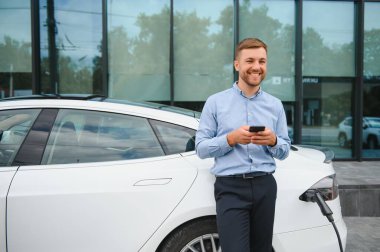 Handsome man in business suit surfing internet on modern smartphone while waiting electric car to charge