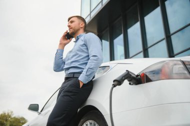 Handsome man in business suit surfing internet on modern smartphone while waiting electric car to charge
