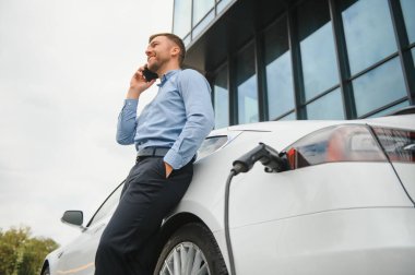 Handsome man in business suit surfing internet on modern smartphone while waiting electric car to charge
