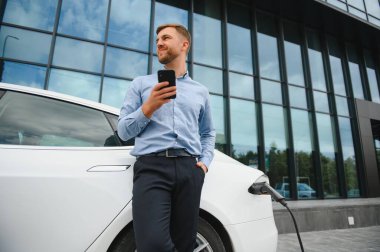 Man charging his electric car at charge station and using smartphone.