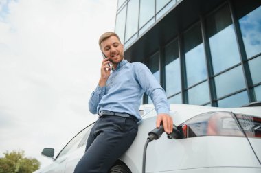 Man charging his electric car at charge station and using smartphone.