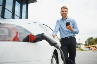 Man charging his electric car at charge station and using smartphone.