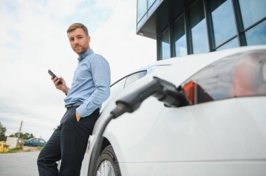 Handsome man in business suit surfing internet on modern smartphone while waiting electric car to charge
