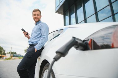 Gayri resmi giyinmiş genç bir adamın portresi, şarj istasyonunda duruyor. Eko elektrikli araba konsepti