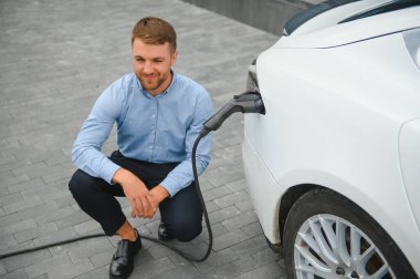 Yakışıklı sakallı adam yeni modern elektrikli arabasının yanında oturuyor ve şarj aletinin fişini çekiyor, araba şarj istasyonunda şarj olurken