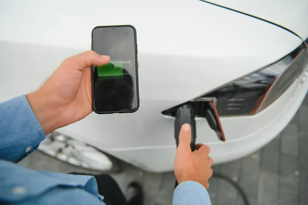 Charging Electric car Looking at App On Mobile Phone. Close up of smartphone screen. Hand holding smart device. Mobile application for eco transportation.