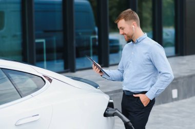 Man charging his electric car at charge station and using smartphone.
