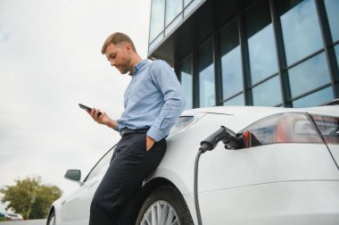 Man charging his electric car at charge station and using smartphone.