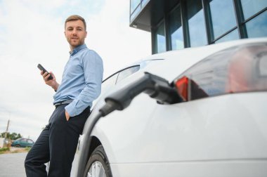 Man charging his electric car at charge station and using smartphone.