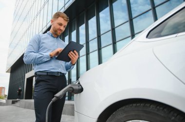 Elektrikli arabanın yanında şarj işleminin bitmesini bekleyen akıllı telefonlu sıradan bir adam..