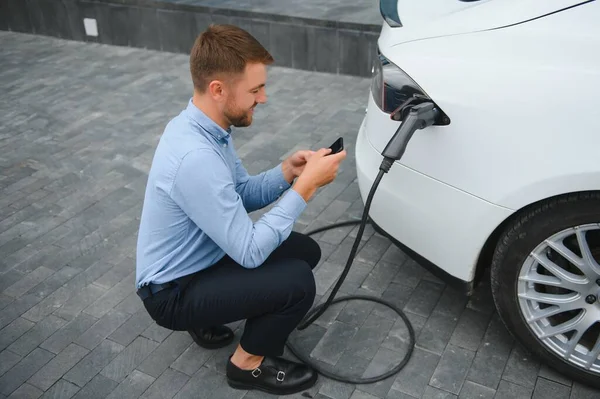 Man charging his electric car at charge station and using smartphone.