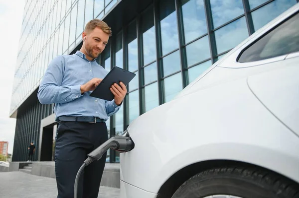 Elektrikli arabanın yanında şarj işleminin bitmesini bekleyen akıllı telefonlu sıradan bir adam..
