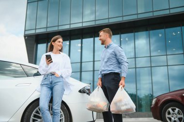 Happy young adult man and smiling woman charging electric car, making shopping and packing bags with food in automobile trunk. Two friends talking and spending free time together.