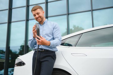 portrait of young handsome bearded man in casual wear, standing at the charging station and holding a plug of the charger for an electric car. Eco electric car concept