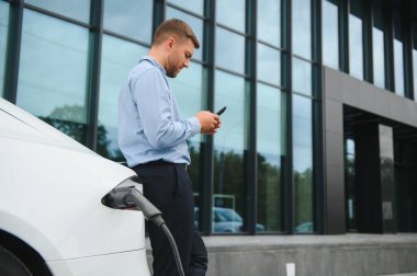 Man charging his electric car at charge station and using smartphone.