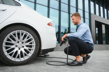 Yakışıklı sakallı adam yeni modern elektrikli arabasının yanında oturuyor ve şarj aletinin fişini çekiyor, araba şarj istasyonunda şarj olurken