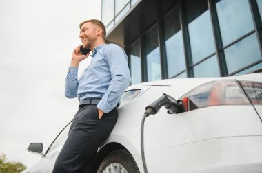 Man charging his electric car at charge station and using smartphone.