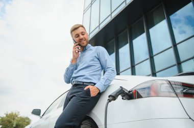 Handsome man in business suit surfing internet on modern smartphone while waiting electric car to charge