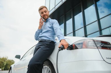 portrait of young handsome bearded man in casual wear, standing at the charging station and holding a plug of the charger for an electric car. Eco electric car concept