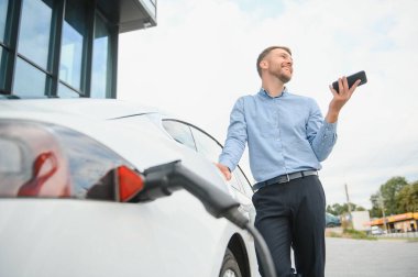 Man charging his electric car at charge station and using smartphone.