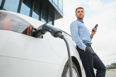 Man charging his electric car at charge station and using smartphone.