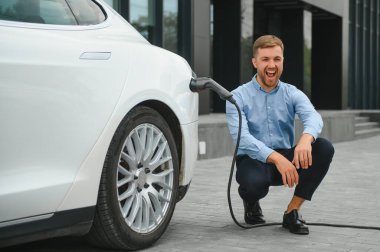 Yakışıklı sakallı adam yeni modern elektrikli arabasının yanında oturuyor ve şarj aletinin fişini çekiyor, araba şarj istasyonunda şarj olurken