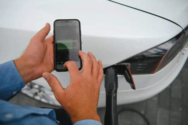 Charging Electric car Looking at App On Mobile Phone. Close up of smartphone screen. Hand holding smart device. Mobile application for eco transportation.