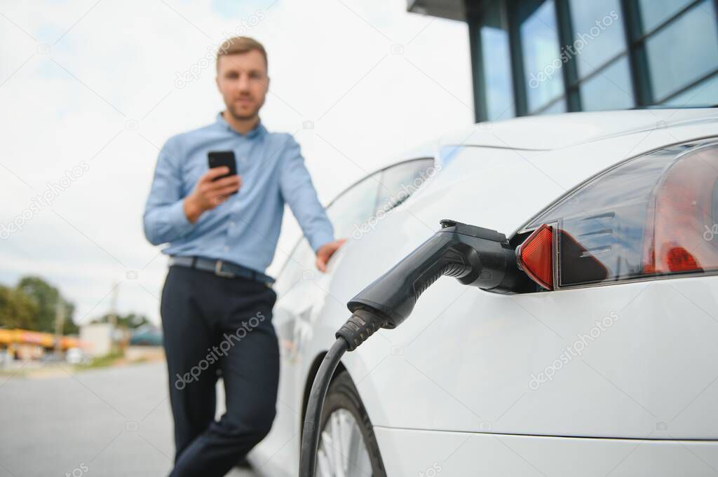 Handsome man in business suit surfing internet on modern smartphone while waiting electric car to charge