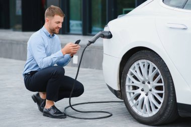 Man charging his electric car at charge station and using smartphone.