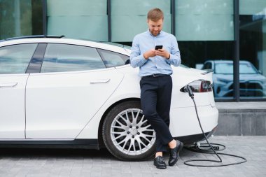 Handsome man in business suit surfing internet on modern smartphone while waiting electric car to charge