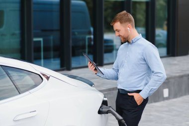 Handsome man in business suit surfing internet on modern smartphone while waiting electric car to charge