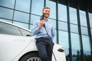 portrait of young handsome bearded man in casual wear, standing at the charging station and holding a plug of the charger for an electric car. Eco electric car concept
