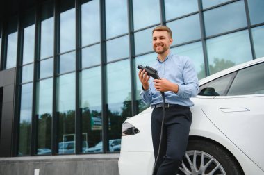 portrait of young handsome bearded man in casual wear, standing at the charging station and holding a plug of the charger for an electric car. Eco electric car concept