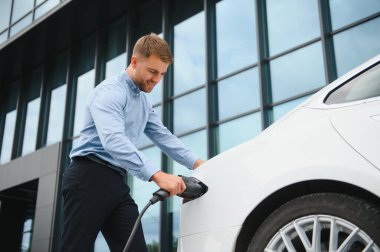 portrait of young handsome bearded man in casual wear, standing at the charging station and holding a plug of the charger for an electric car. Eco electric car concept