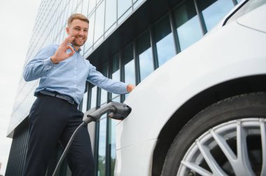 Gayri resmi giyinmiş genç bir adamın portresi, şarj istasyonunda duruyor. Eko elektrikli araba konsepti