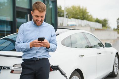 Man charging his electric car at charge station and using smartphone.