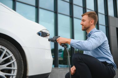 Yakışıklı sakallı adam yeni modern elektrikli arabasının yanında oturuyor ve şarj aletinin fişini çekiyor, araba şarj istasyonunda şarj olurken