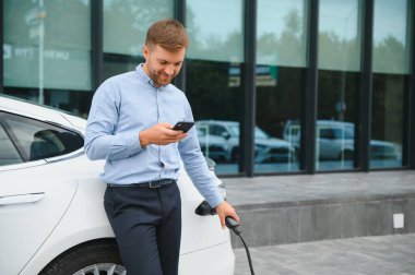 Man charging his electric car at charge station and using smartphone.
