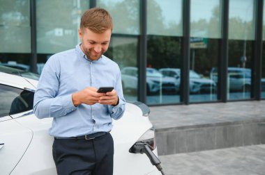 Man charging his electric car at charge station and using smartphone.
