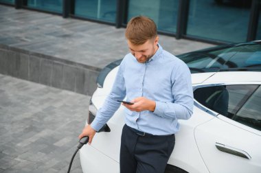 Handsome man in business suit surfing internet on modern smartphone while waiting electric car to charge