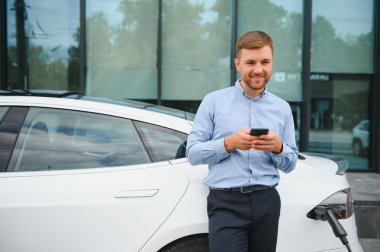 Man charging his electric car at charge station and using smartphone.