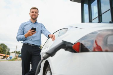 Gayri resmi giyinmiş genç bir adamın portresi, şarj istasyonunda duruyor. Eko elektrikli araba konsepti