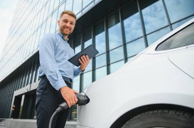 portrait of young handsome bearded man in casual wear, standing at the charging station and holding a plug of the charger for an electric car. Eco electric car concept