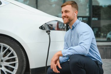 Gayri resmi giyinmiş genç bir adamın portresi, şarj istasyonunda duruyor. Eko elektrikli araba konsepti