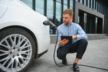 Yakışıklı sakallı adam yeni modern elektrikli arabasının yanında oturuyor ve şarj aletinin fişini çekiyor, araba şarj istasyonunda şarj olurken