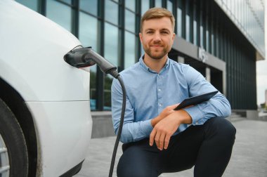 Yakışıklı sakallı adam yeni modern elektrikli arabasının yanında oturuyor ve şarj aletinin fişini çekiyor, araba şarj istasyonunda şarj olurken